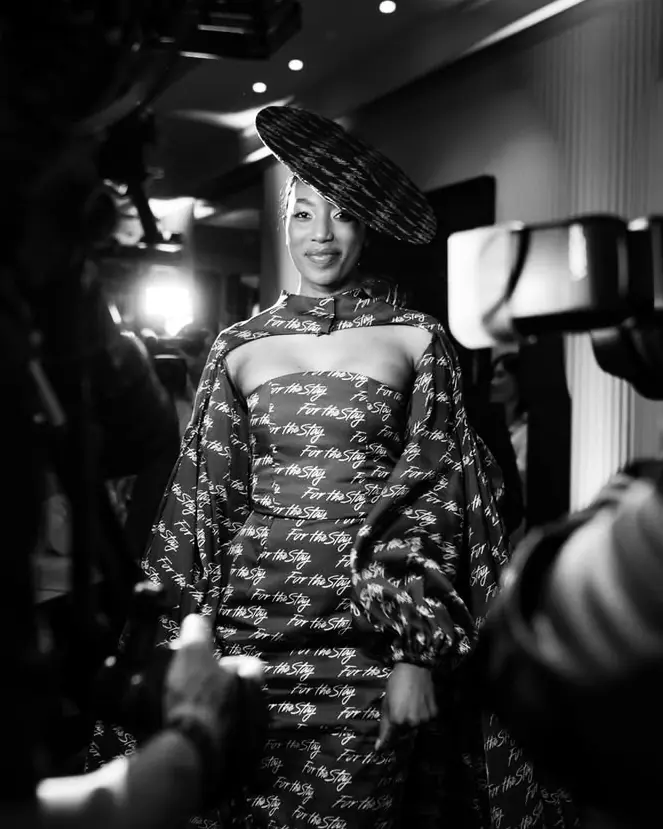 The finished dress worn with a wide-brimmed hat, photographers at the edge of the frame.