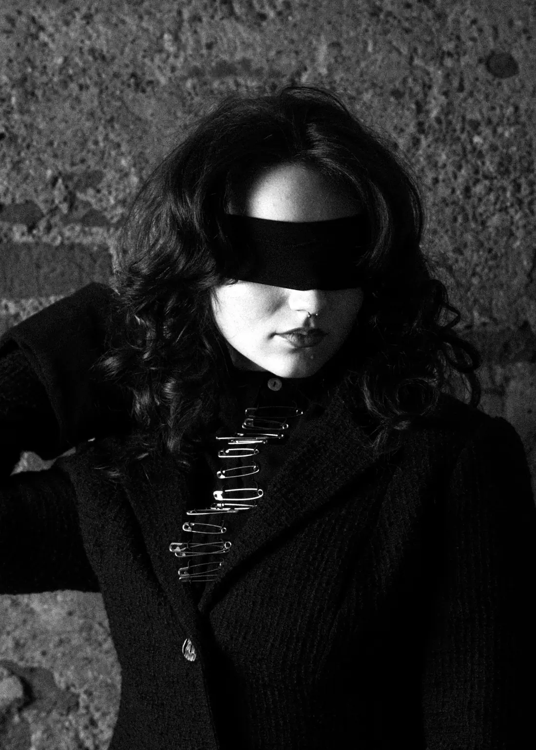 Portrait in a black knit jacket fastened with a row of safety pins, eyes covered by a black band, against a concrete wall.
