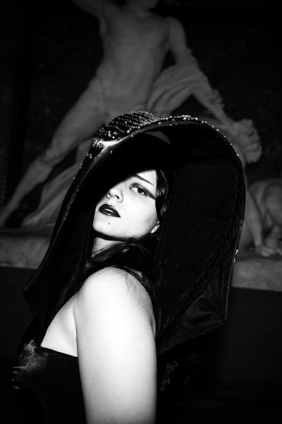 Close portrait under the wide black brim of the headpiece, a marble sculpture behind.